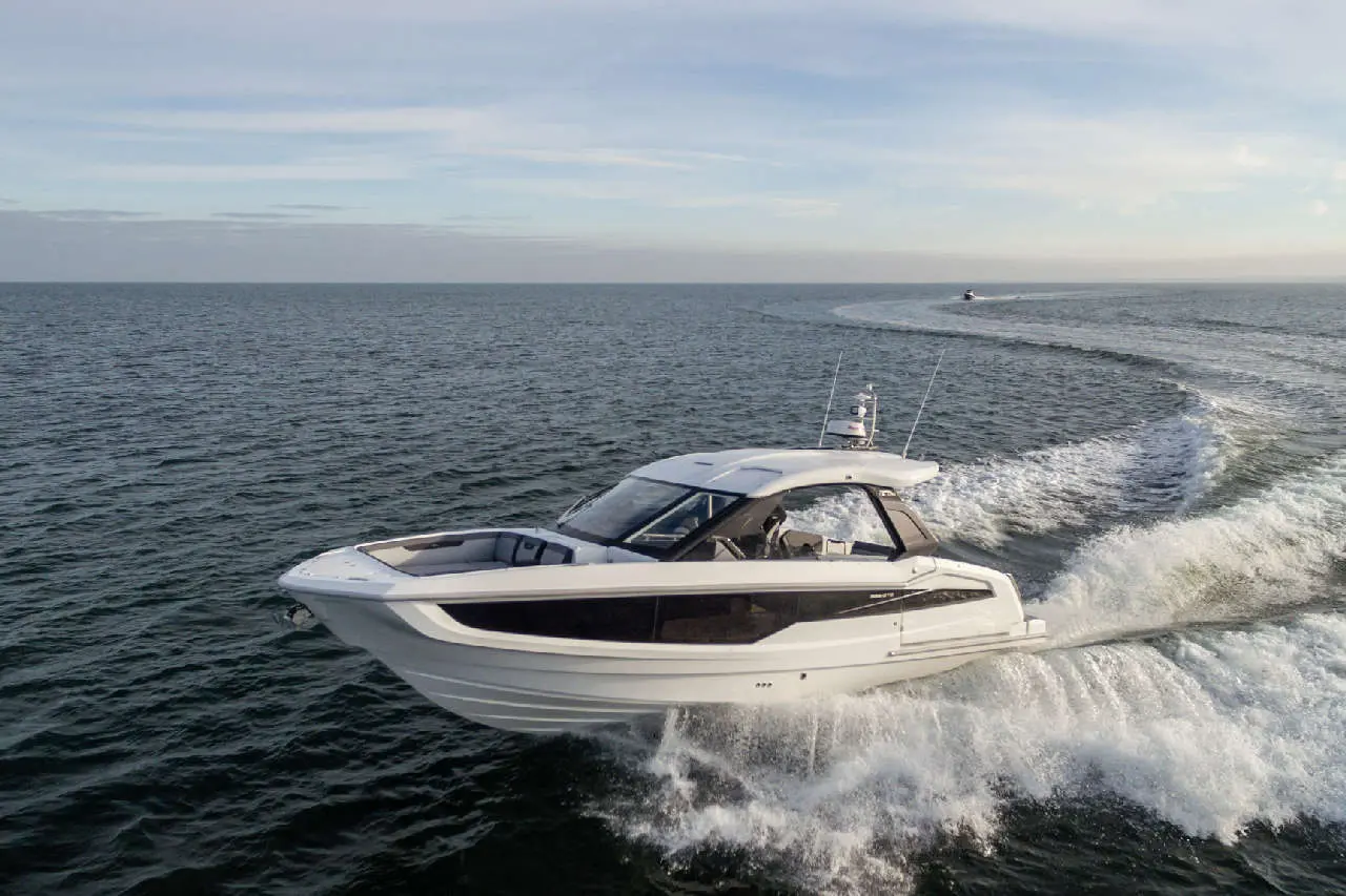 A sleek white motor yacht glides over the water, creating waves, under a clear sky.