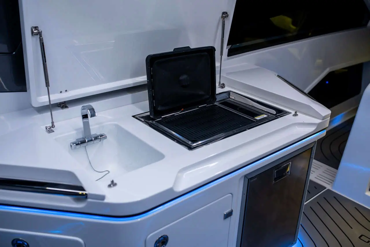 A modern boat's outdoor kitchen with a sink, grill, and storage, featuring sleek white surfaces and blue LED lighting.