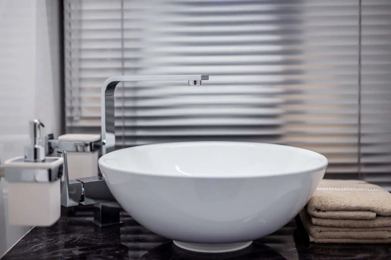Sleek modern bathroom sink, chrome tap, and folded towels beside a white bowl sink against a textured blind backdrop.