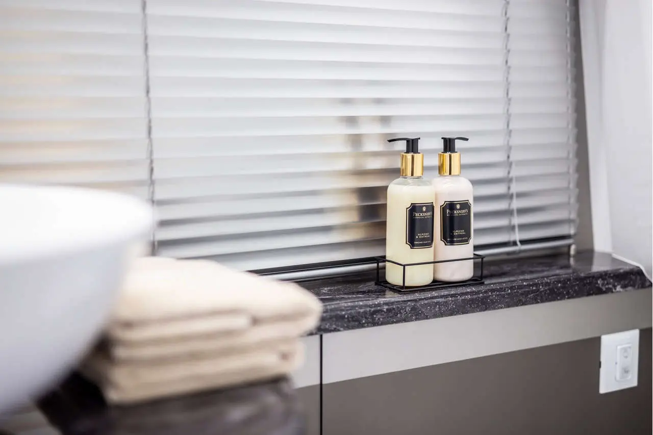 Two elegant shampoo and conditioner bottles on a marble shelf, beside folded towels, with blinds in the background.
