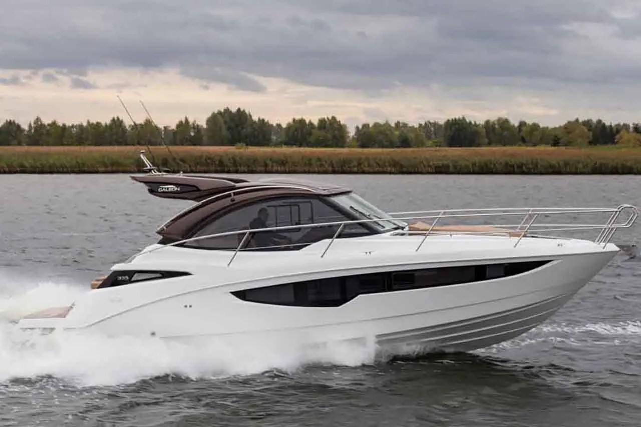 A sleek white motorboat steams across a calm river, surrounded by lush greenery under a cloudy sky.