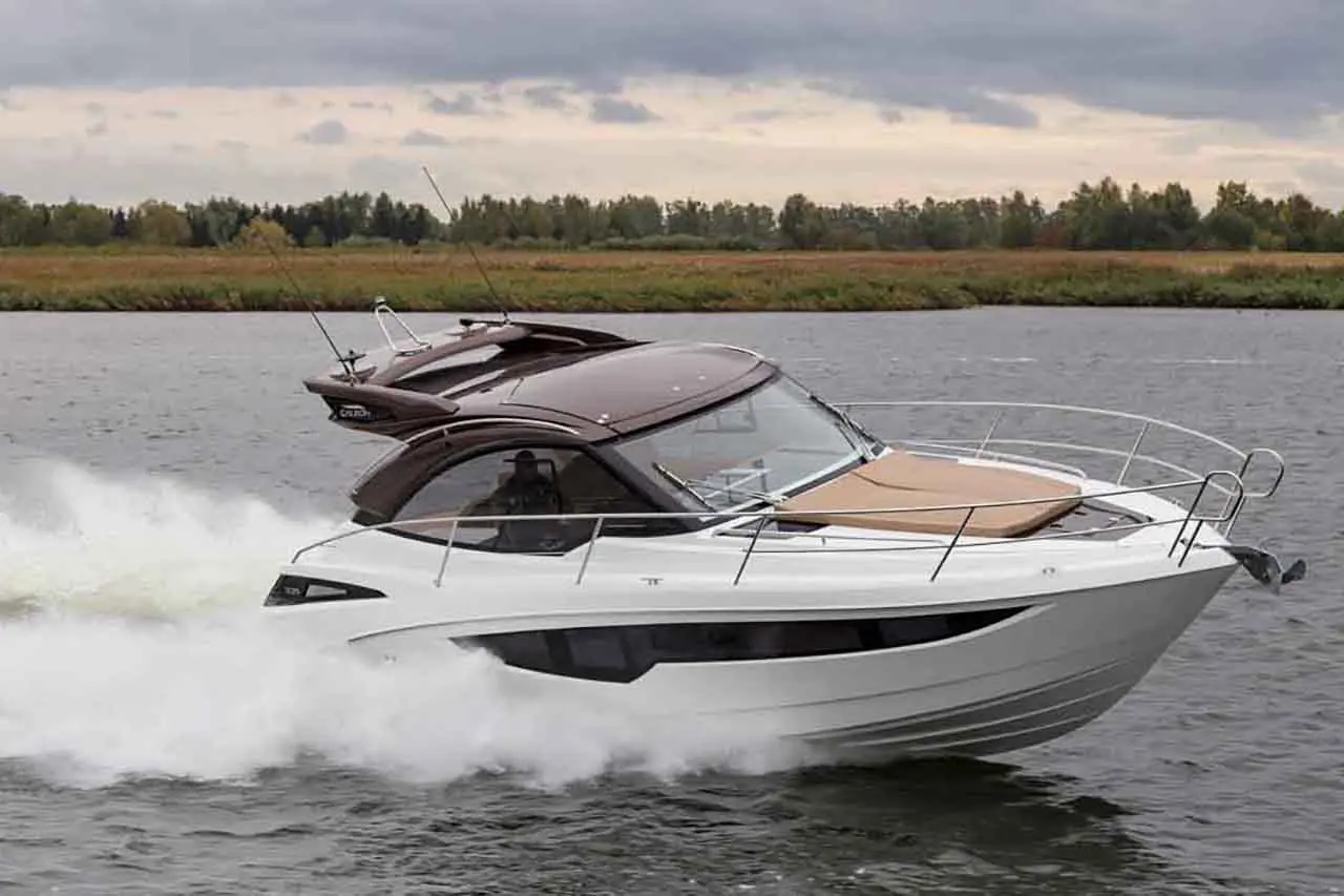 A sleek white motorboat speeds across a calm river, leaving a wake, with green trees lining the shore in the background.