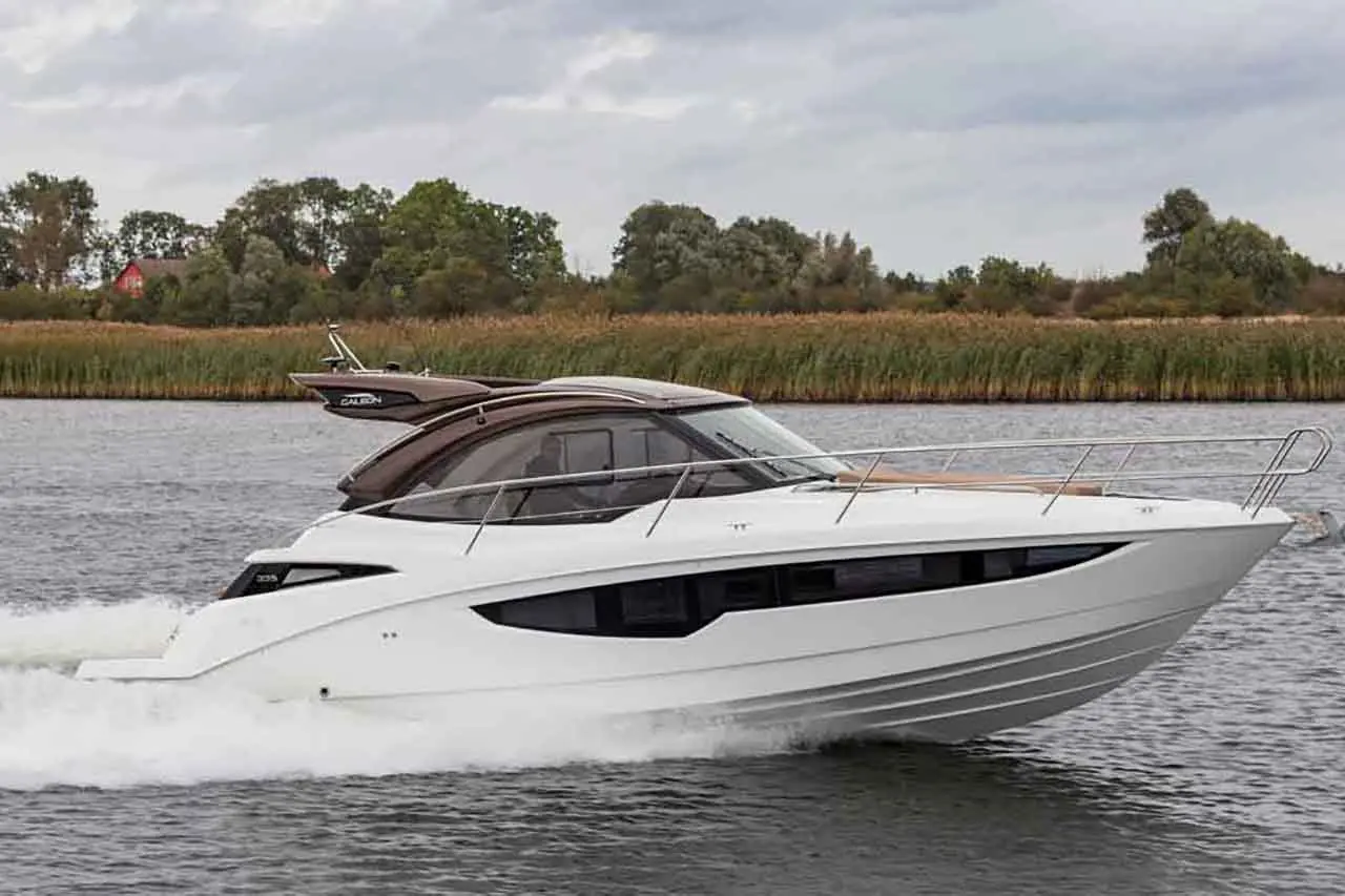 A sleek white motorboat speeds through a calm river, surrounded by lush greenery and an overcast sky.
