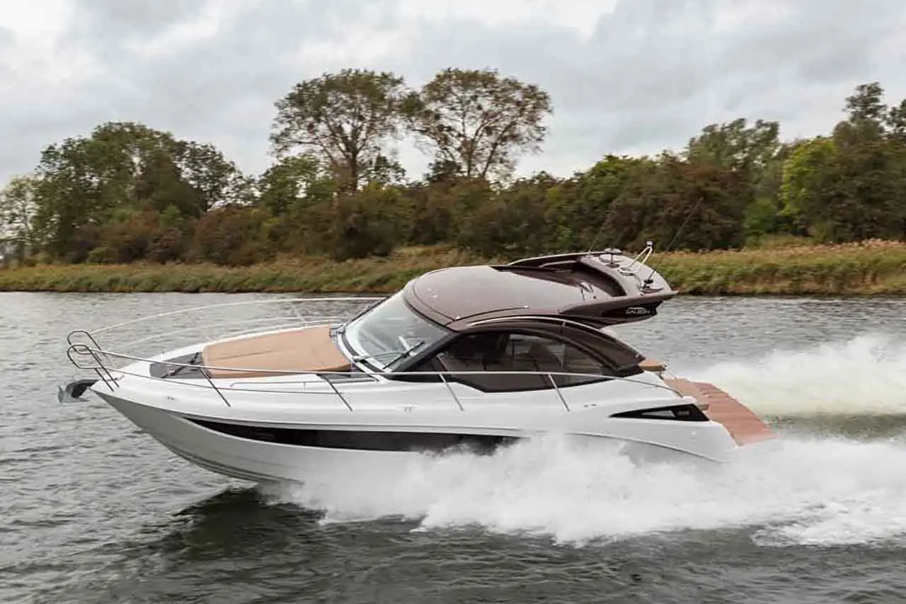 A sleek white motorboat speeds across a river, leaving a wake, with lush greenery in the background under a cloudy sky.