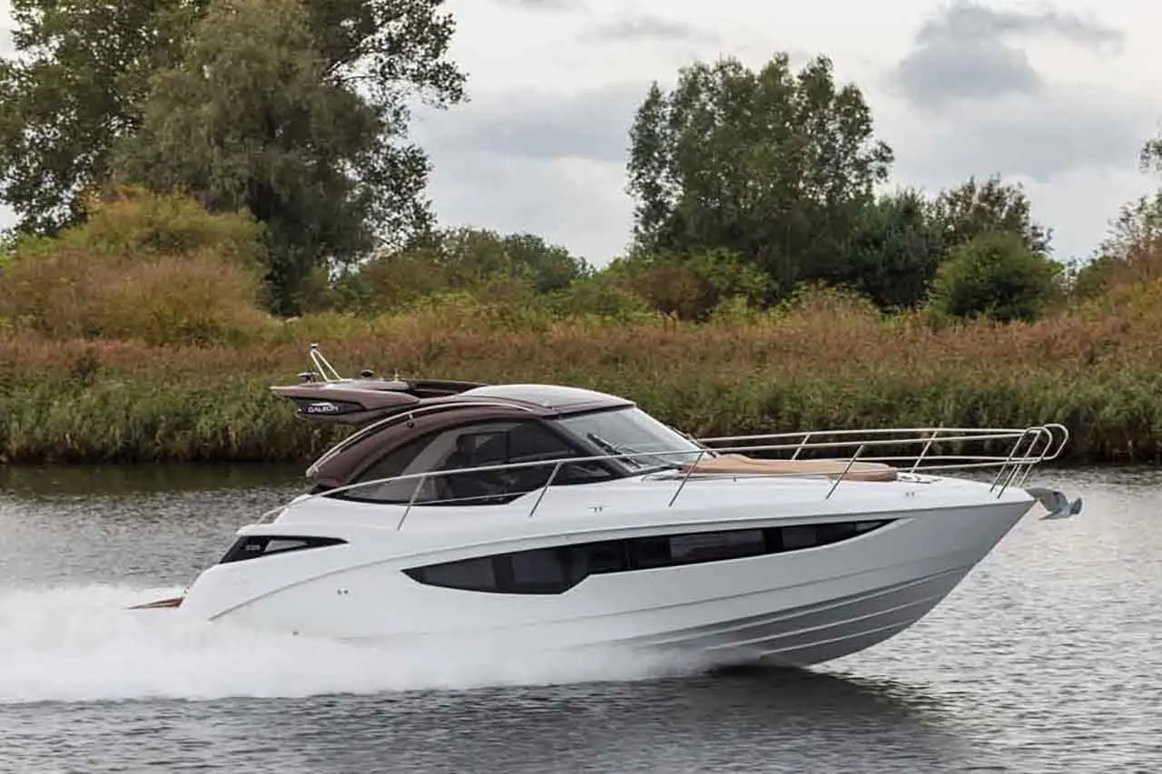 A sleek white motorboat speeds along a river, surrounded by lush greenery and cloudy skies.