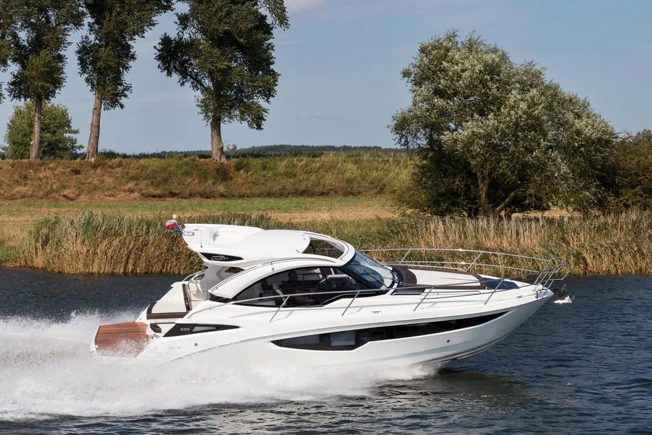 A sleek white motorboat glides across calm waters, with tall trees and grassy banks in the background under a clear blue sky.