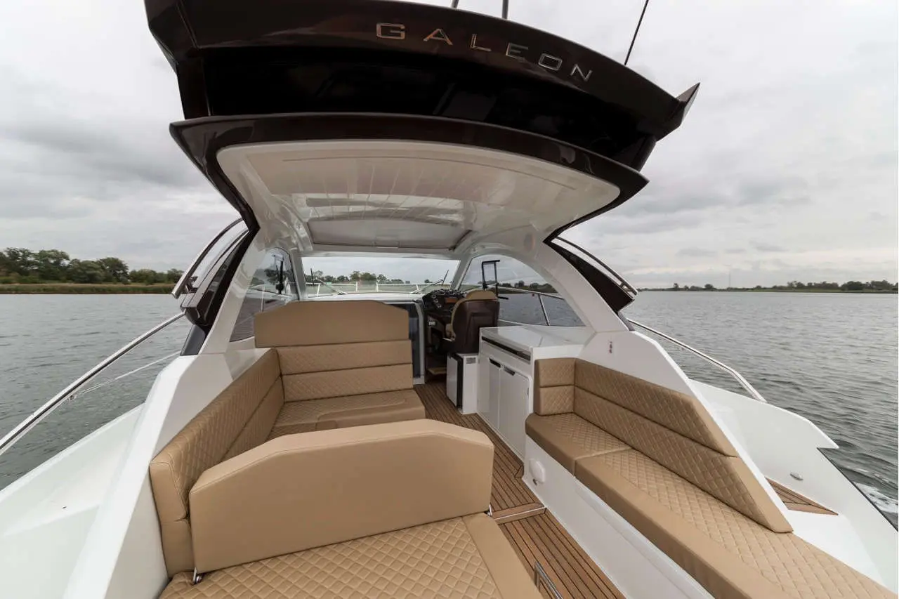 A view of a luxurious boat interior, featuring tan seating and a helm, with a scenic water backdrop under cloudy skies.