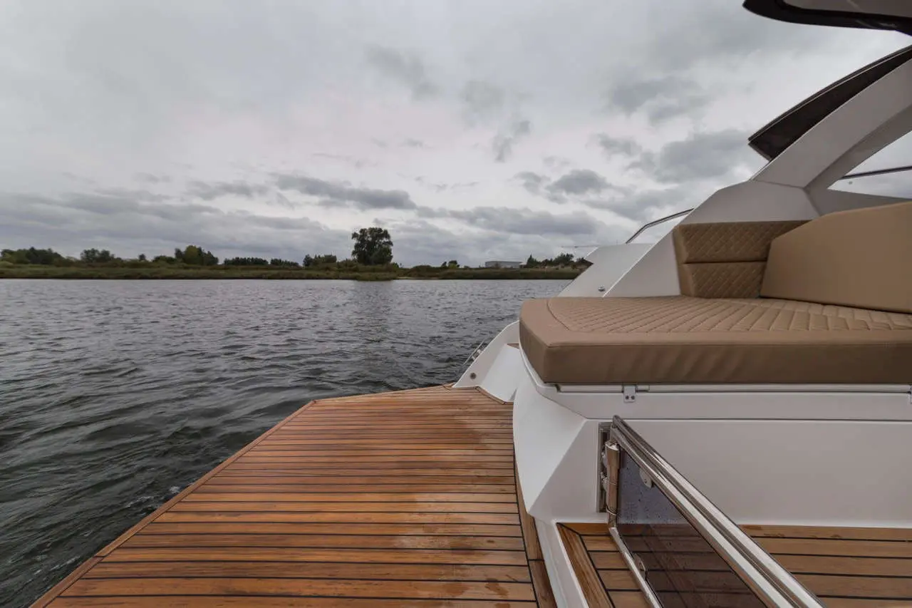 A comfortable boat with a beige seat is docked by a wooden platform on a calm body of water under a cloudy sky.