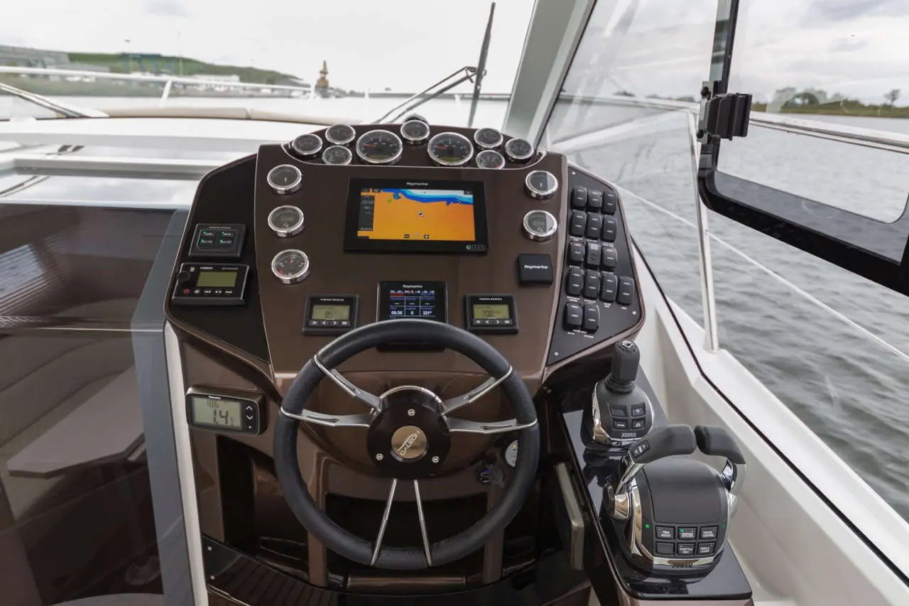 Boat cockpit with steering wheel, navigation screen, gauges, and controls overlooking calm waters, featuring visible depth readings.