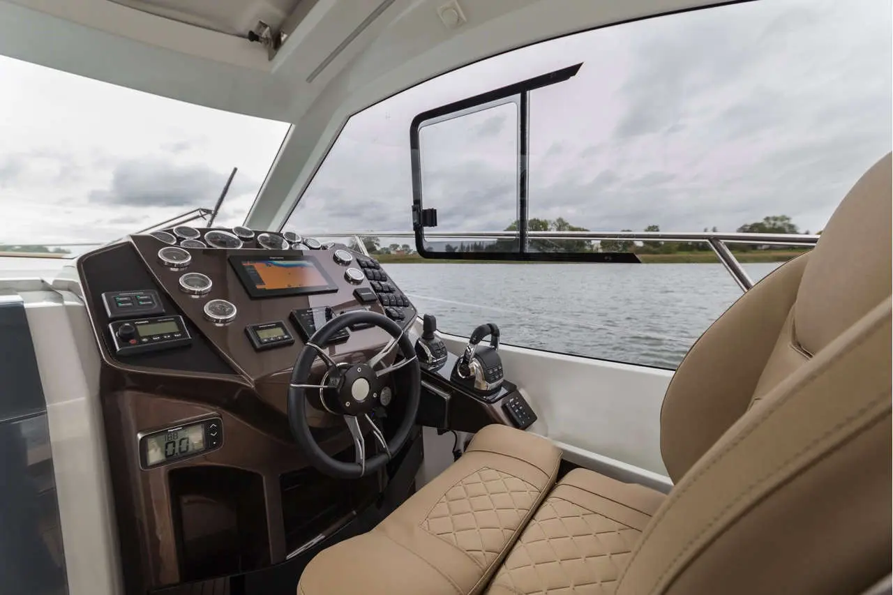Modern boat dashboard with steering wheel, control panel, and digital screen, overlooking a calm waterway and cloudy sky.