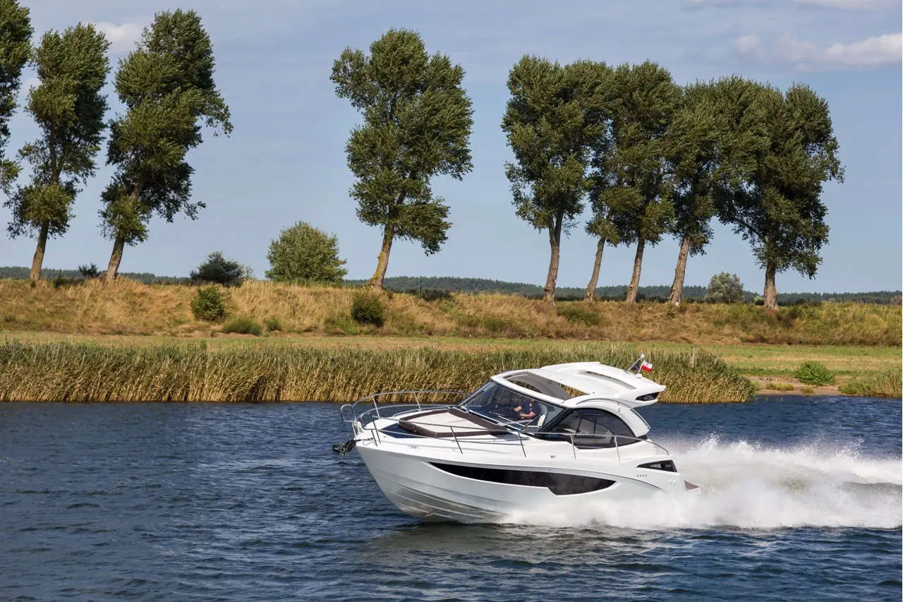 A white motorboat speeds across a serene river, flanked by green trees and lush banks under a clear blue sky.