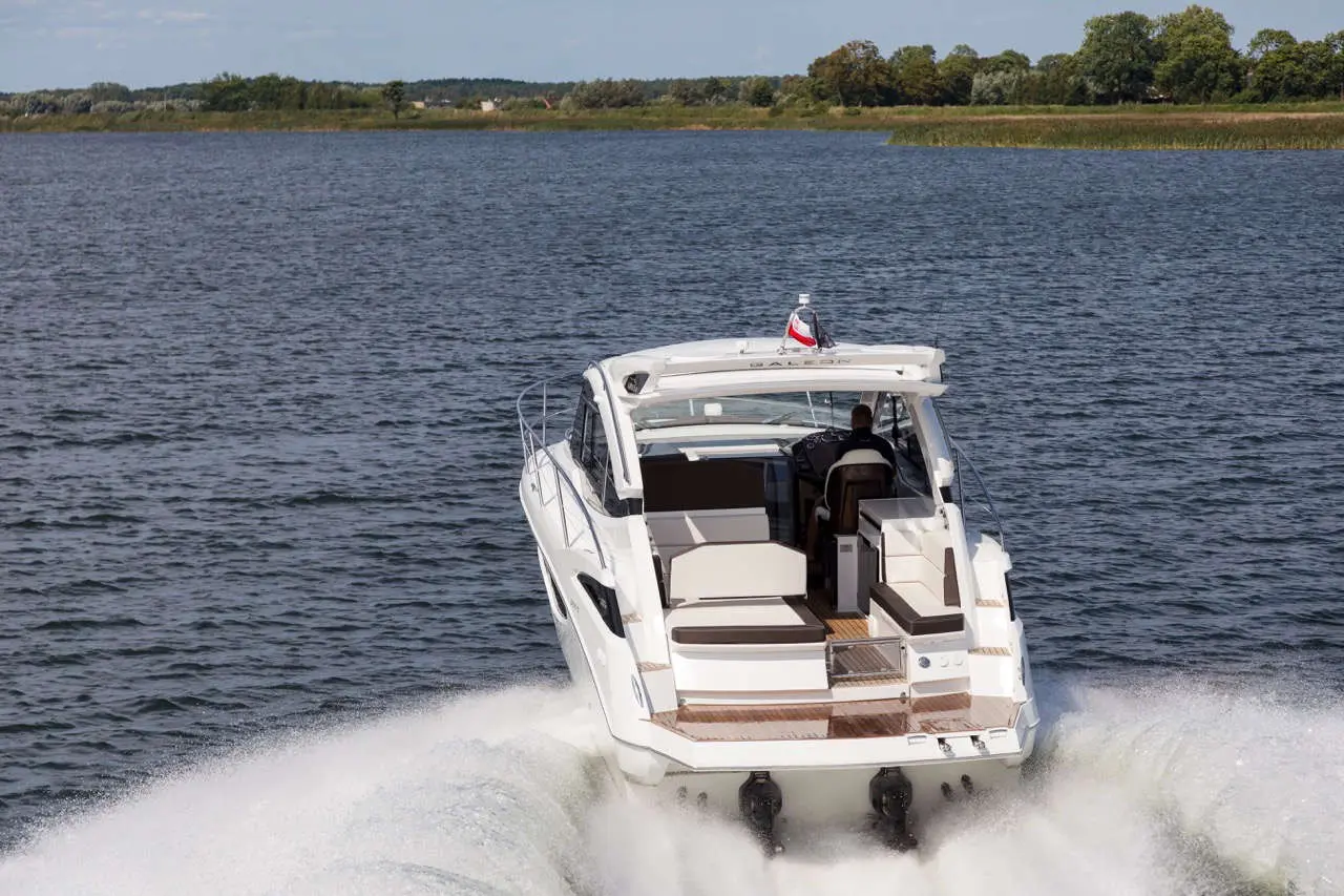 A motorboat speeding on a calm waterway, with a driver visible at the helm and a scenic landscape in the background.