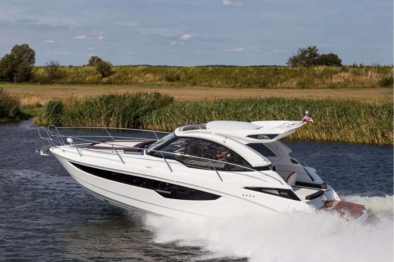 A sleek white motorboat with a modern design speeds through calm water, surrounded by lush greenery and a clear sky.