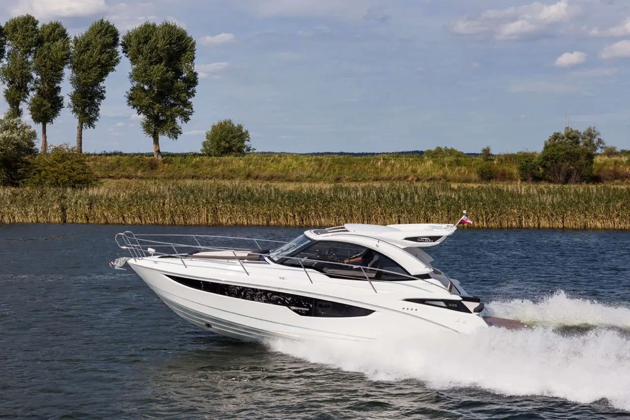 A sleek white motorboat glides through calm waters, surrounded by lush greenery and tall trees under a blue sky.