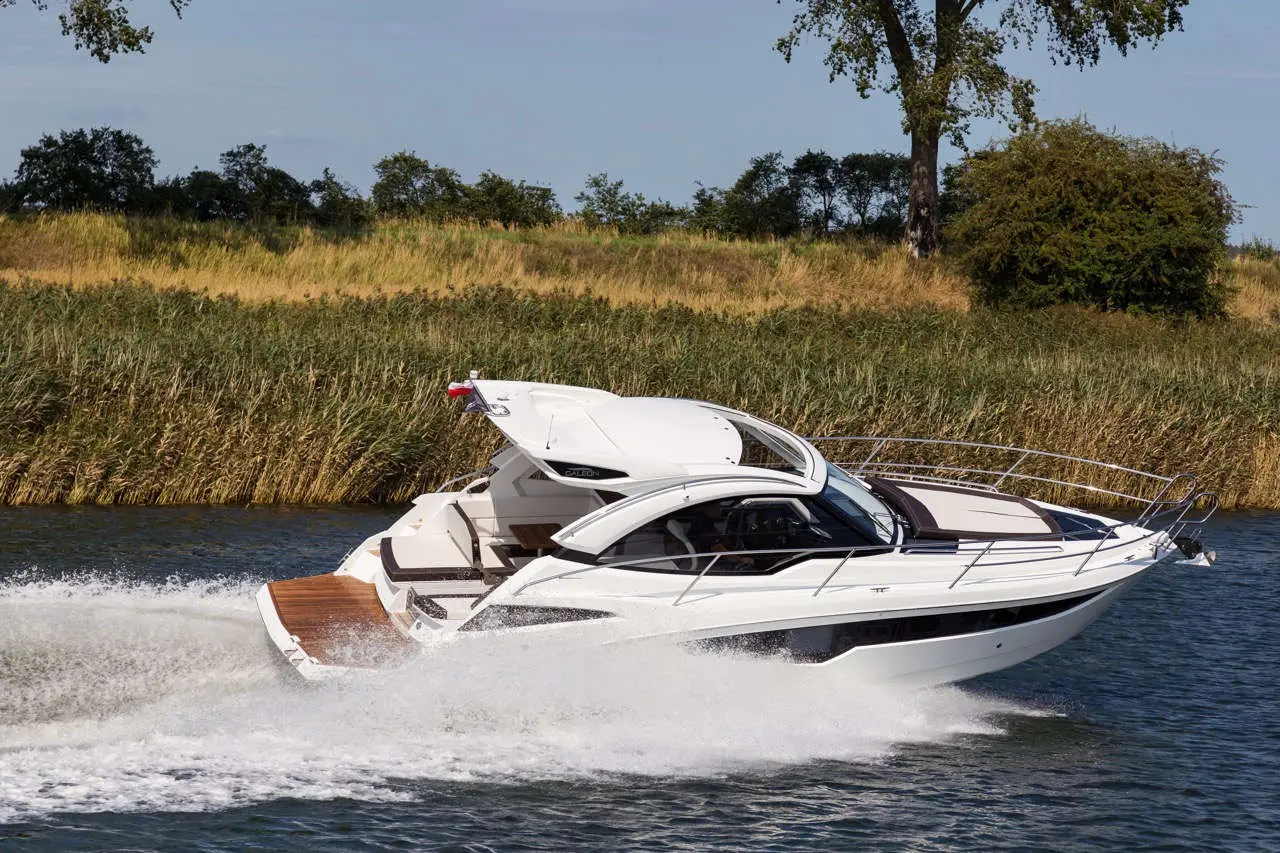 A stylish white motorboat speeds across the water, leaving a wake, with lush greenery lining the shoreline in the background.