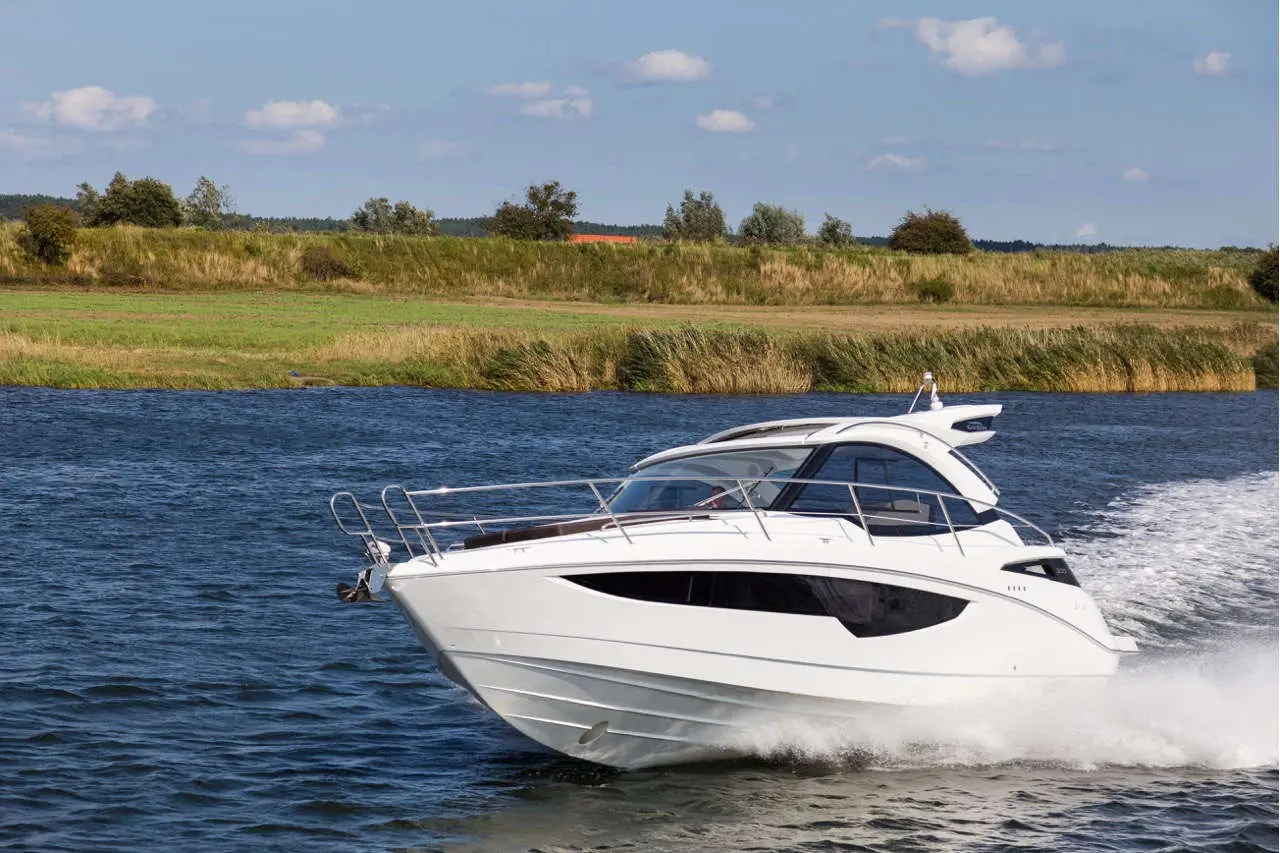 A sleek white motorboat speeds through water, surrounded by green grass and trees under a bright blue sky.