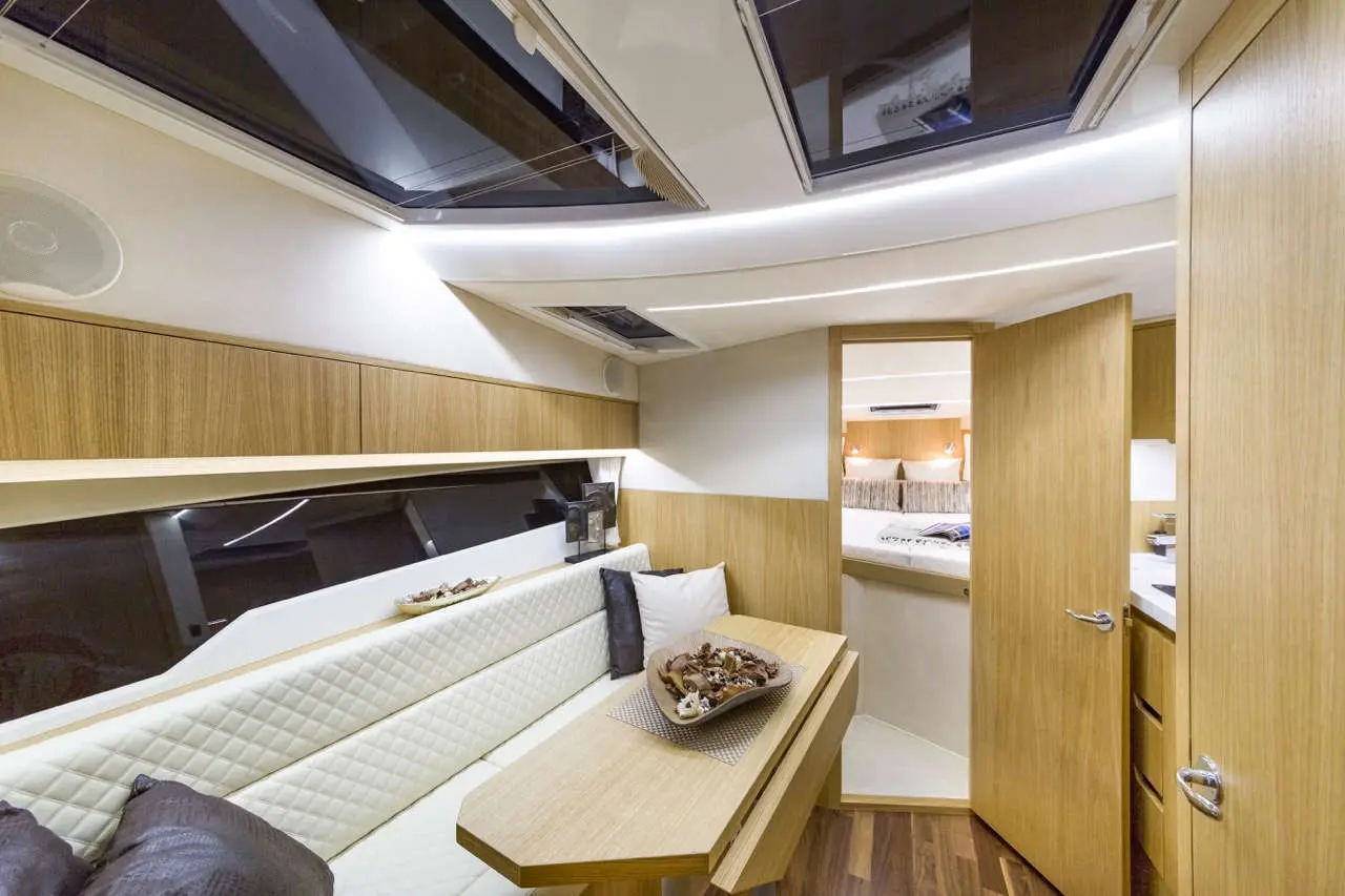 Modern yacht interior featuring a light wood finish, seating area with cushions, and an open door leading to the bedroom.