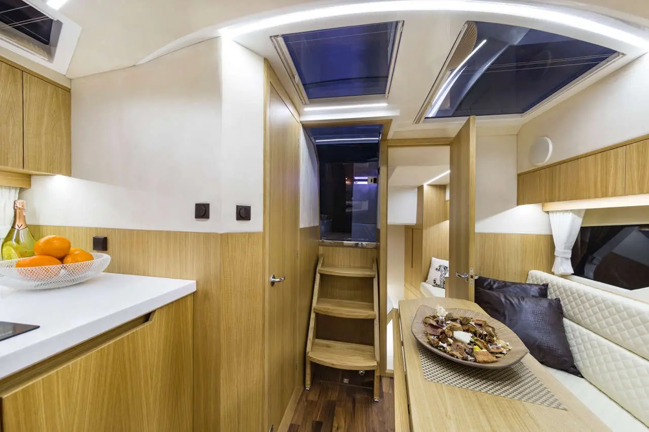 Modern boat interior showing a cozy galley with a white countertop, fruit bowl, and wooden stairs leading to a living area.