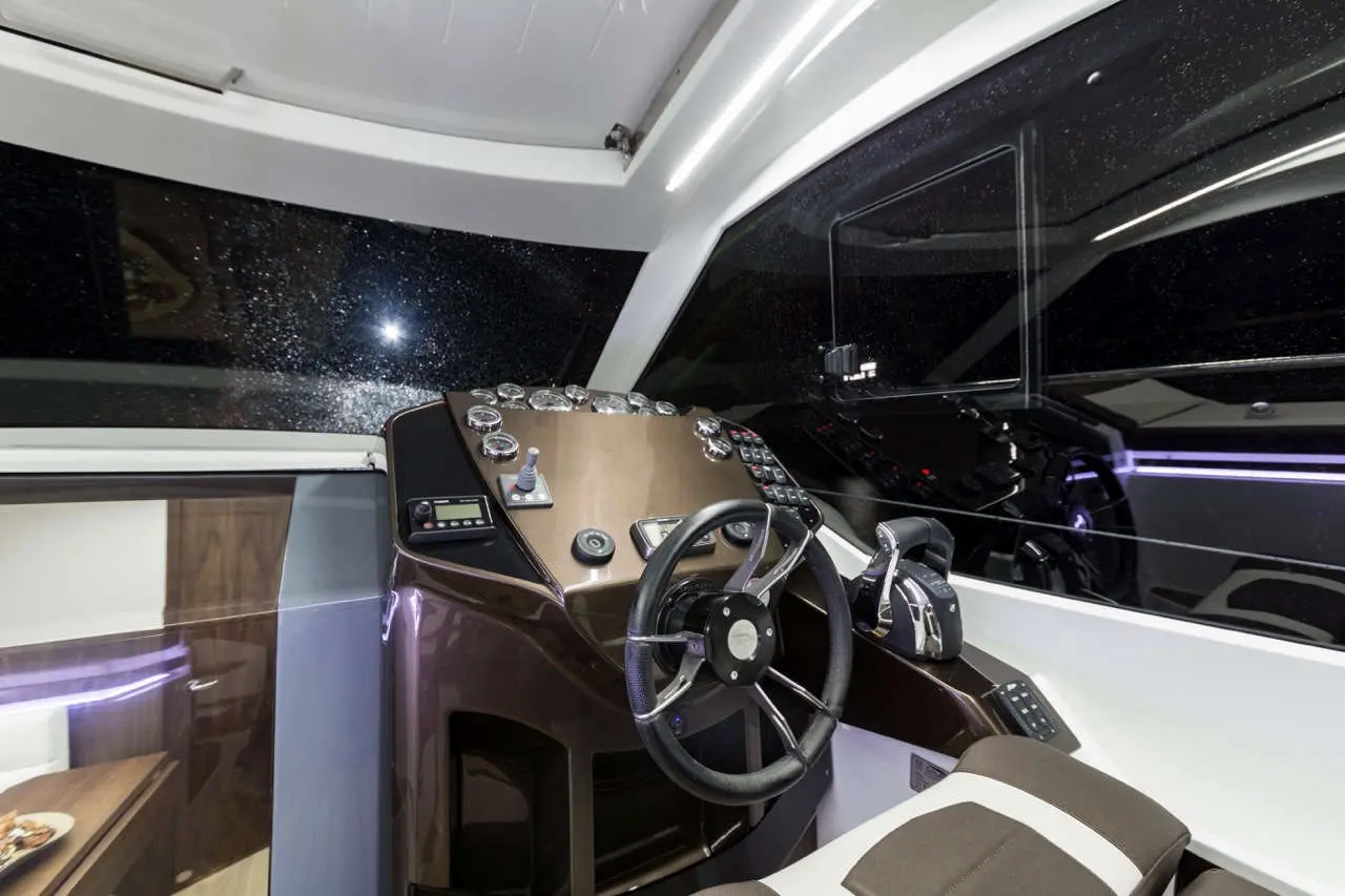 Modern yacht cockpit featuring steering wheel, gauges, and control panels, with a view through rain-speckled glass.