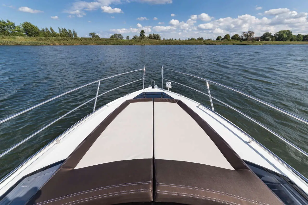 Bow of a boat with sunbathing cushions, overlooking serene water and lush greenery under a blue sky with fluffy clouds.