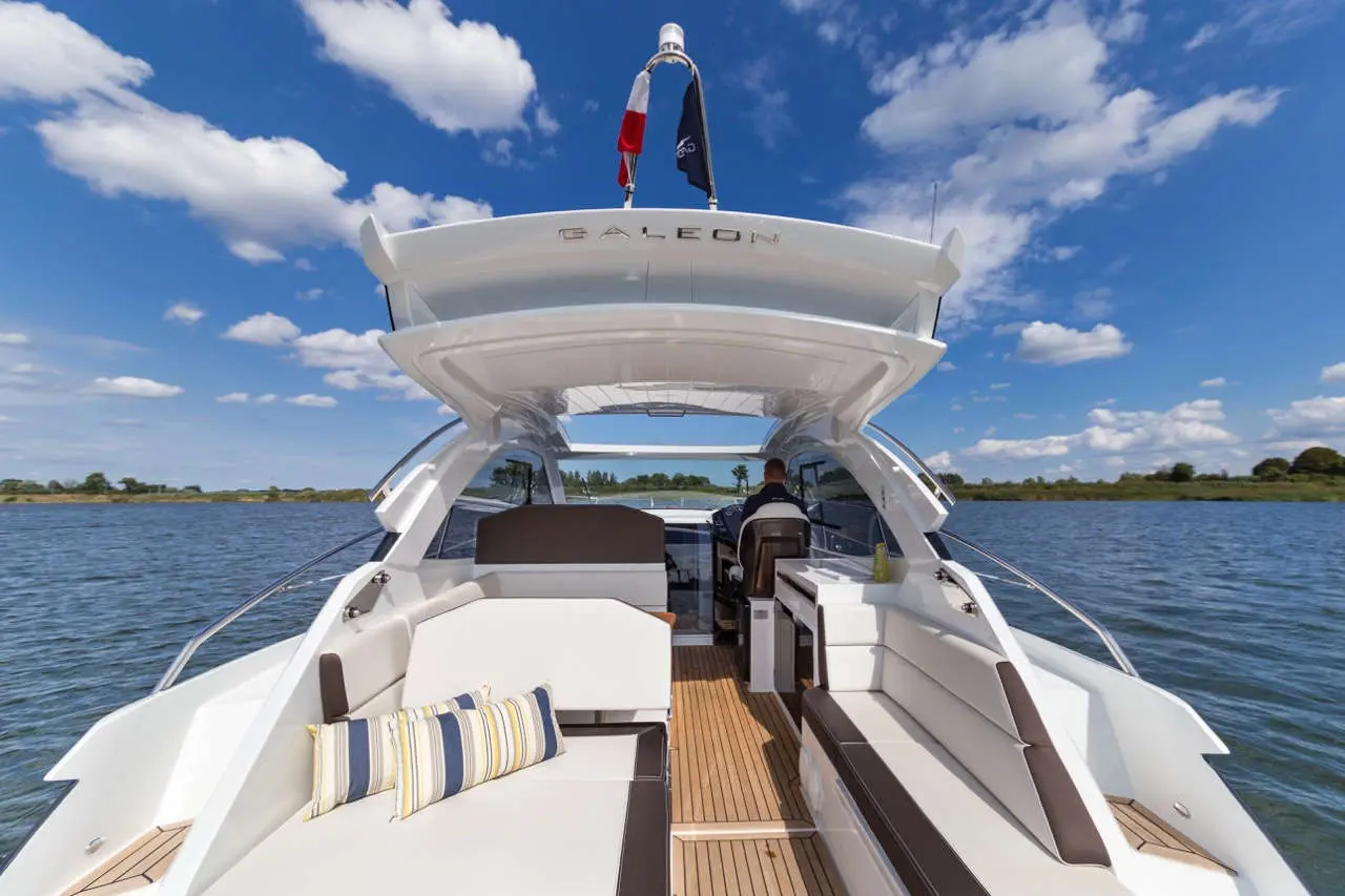 A sleek Galeon boat interior with a captain at the helm, plush seating, and a scenic water view under a blue sky.