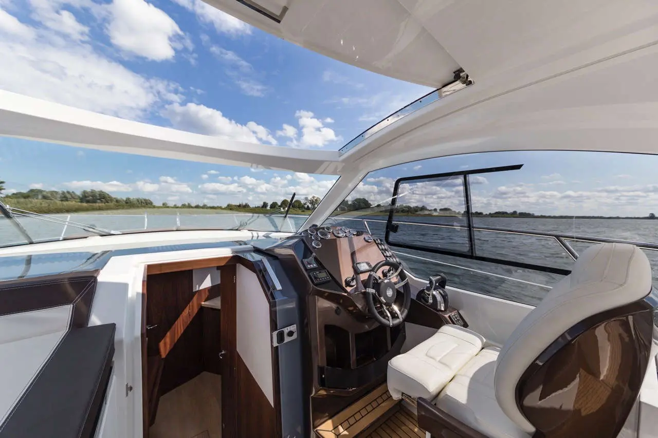Interior view of a luxury boat cockpit showing steering wheel, dashboard, and river scenery through large windows.