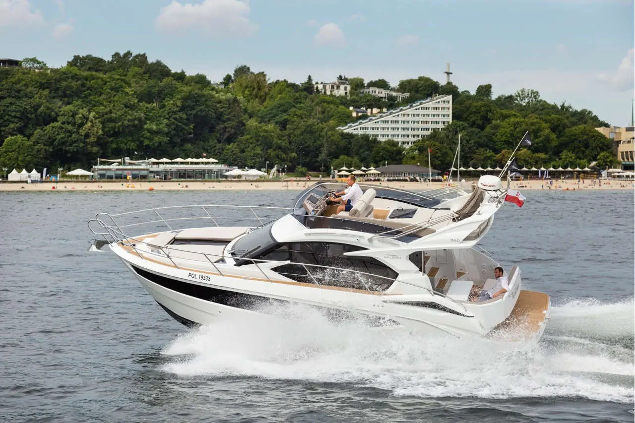 A white luxury boat with the registration POL 19333 navigates smoothly on water near a beach and green hillside.