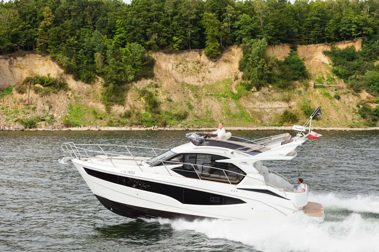 A white luxury motorboat cruising on water near a green cliff, with two people onboard, showcasing leisure and adventure.