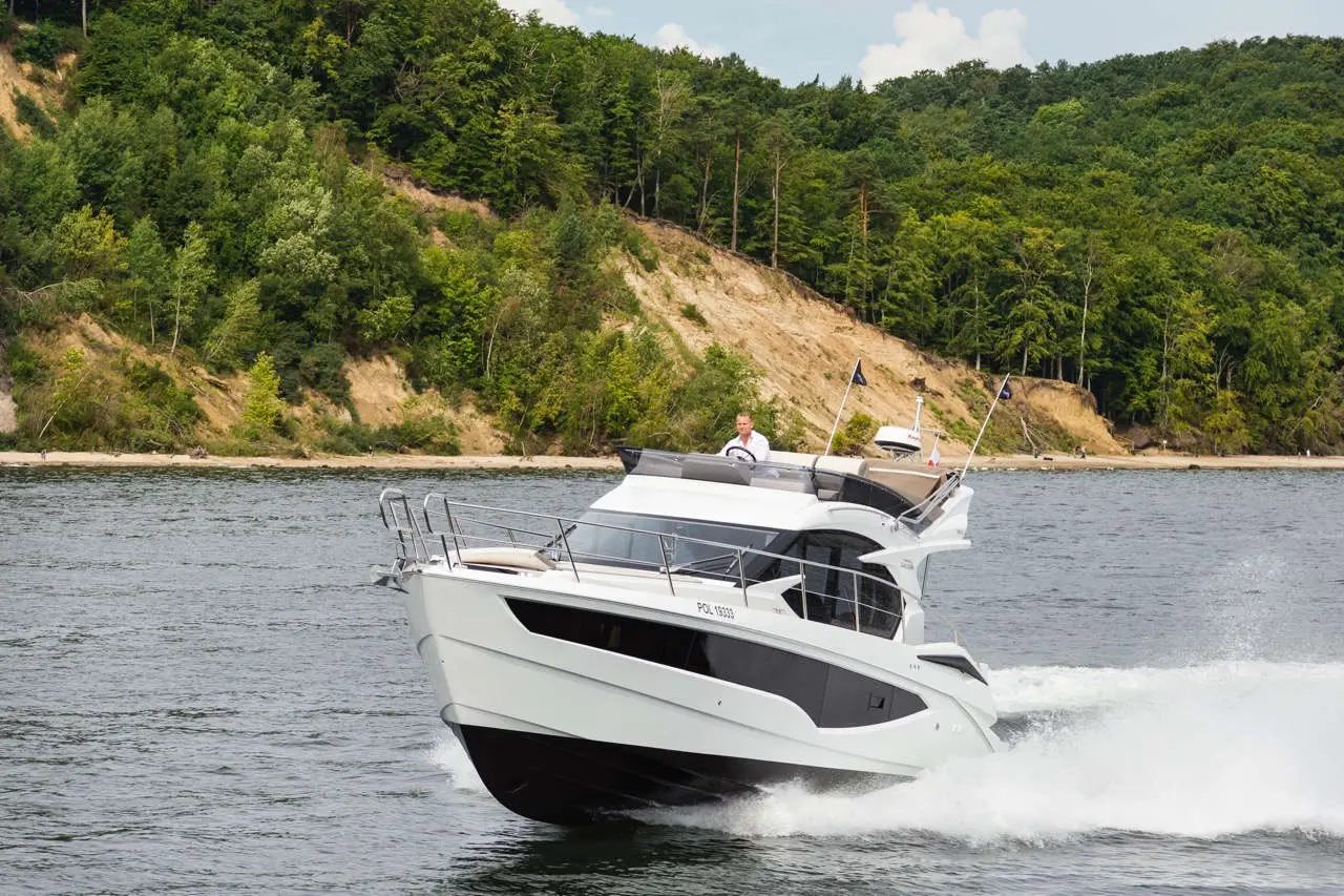 A sleek white motorboat speeds across a lake with a forested hillside in the background; a man is at the helm.