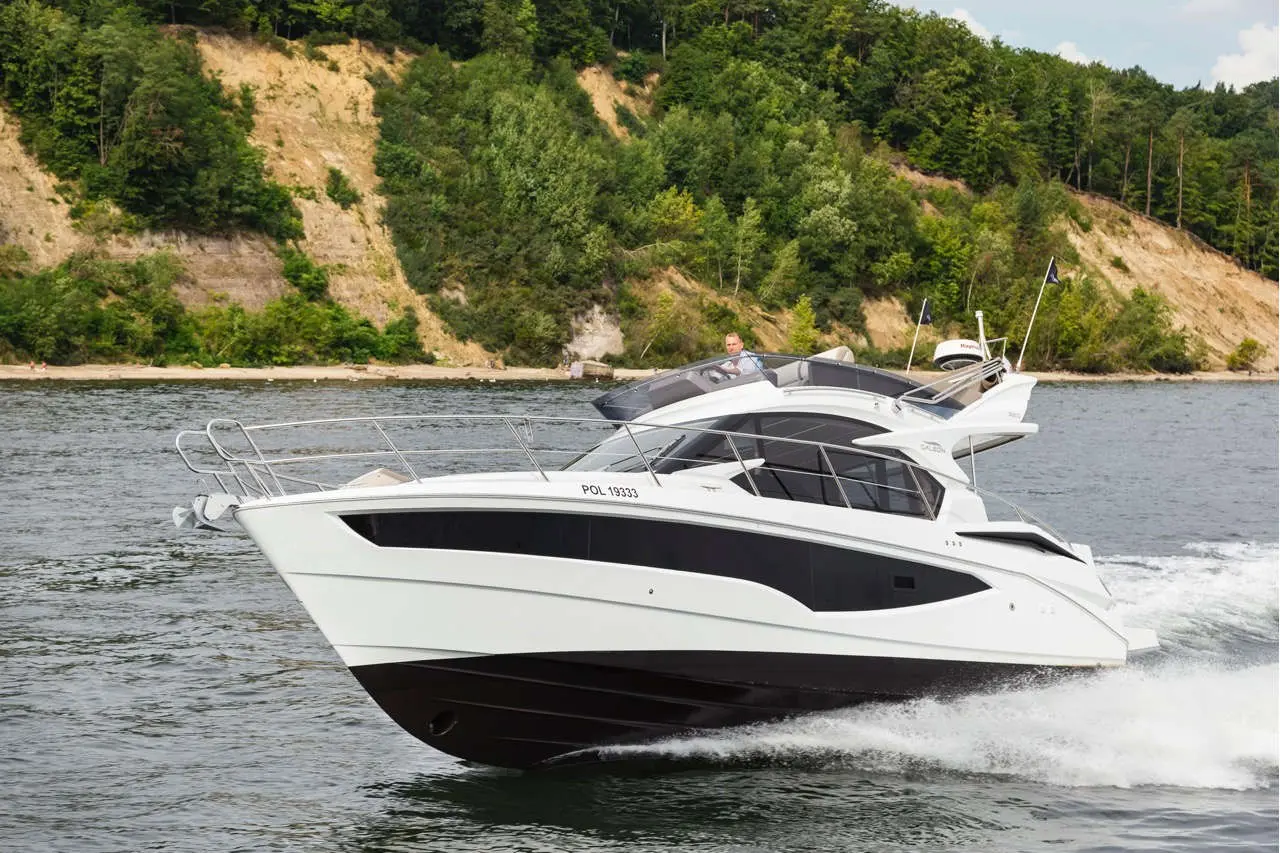 A white and black luxury yacht with the registration POL 19333 glides through a river, surrounded by lush greenery.