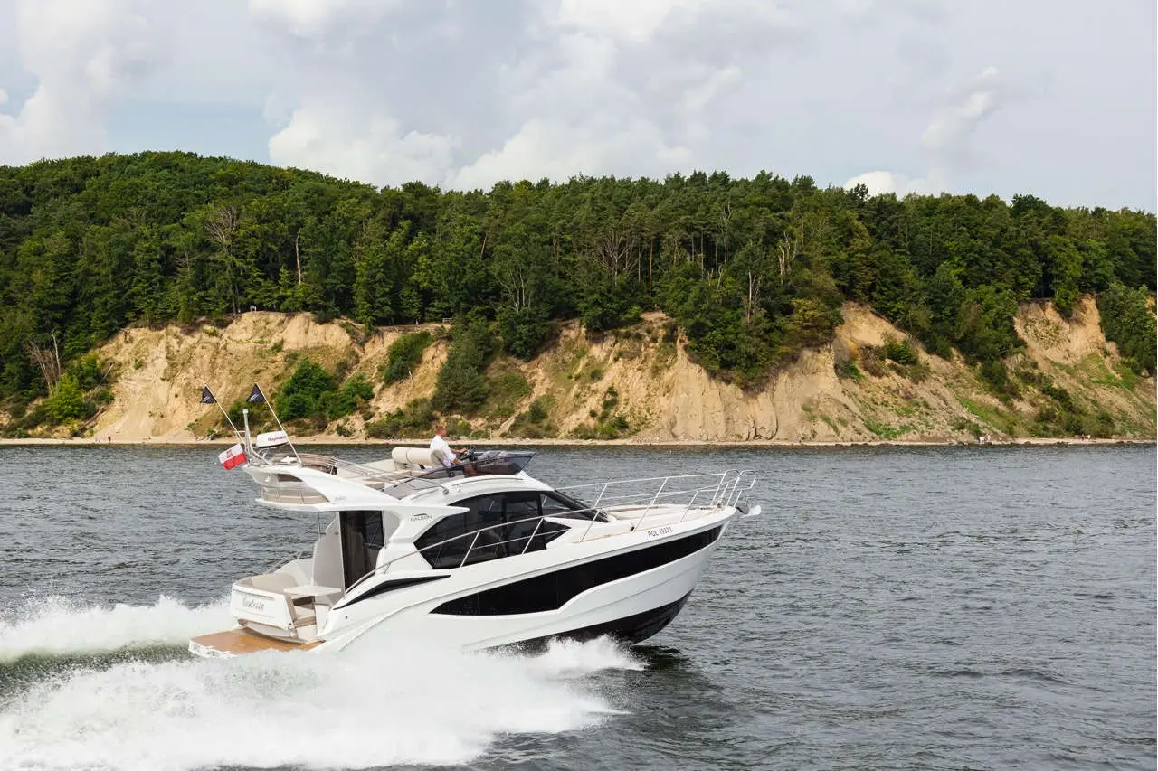 A motorboat cruises through calm waters near a wooded hillside, showcasing flags and sleek design against a scenic backdrop.