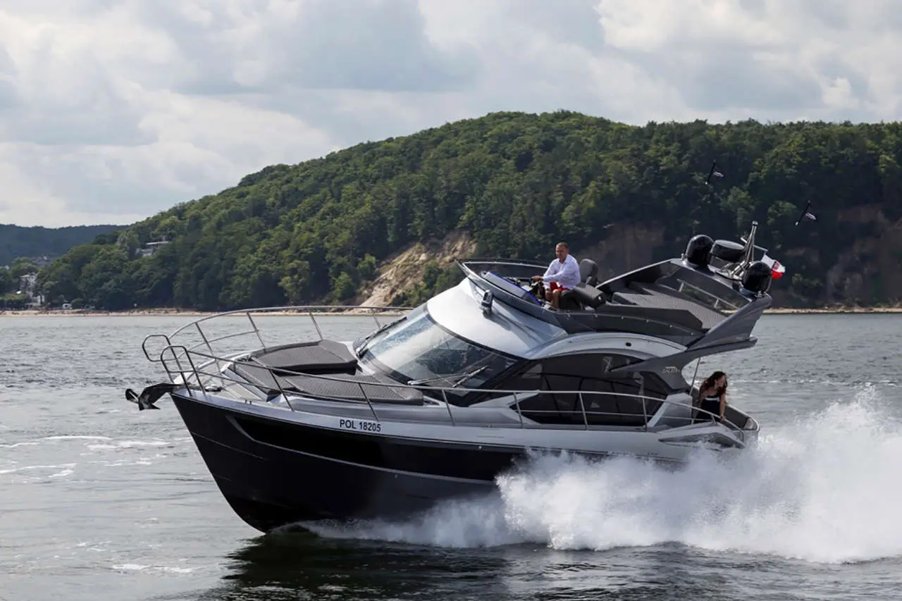 A black and silver motorboat speeds through water, creating splashes, with two people onboard against a forested backdrop.