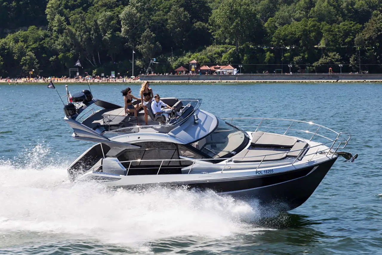 A sleek motorboat speeds across the water with people on board, set against a lush green shoreline and a crowded beach.