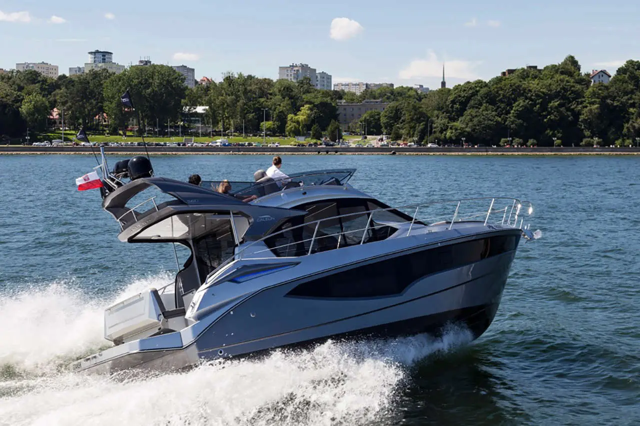 A sleek gray motor yacht speeds across the water, with a Polish flag flying and city skyline in the background.