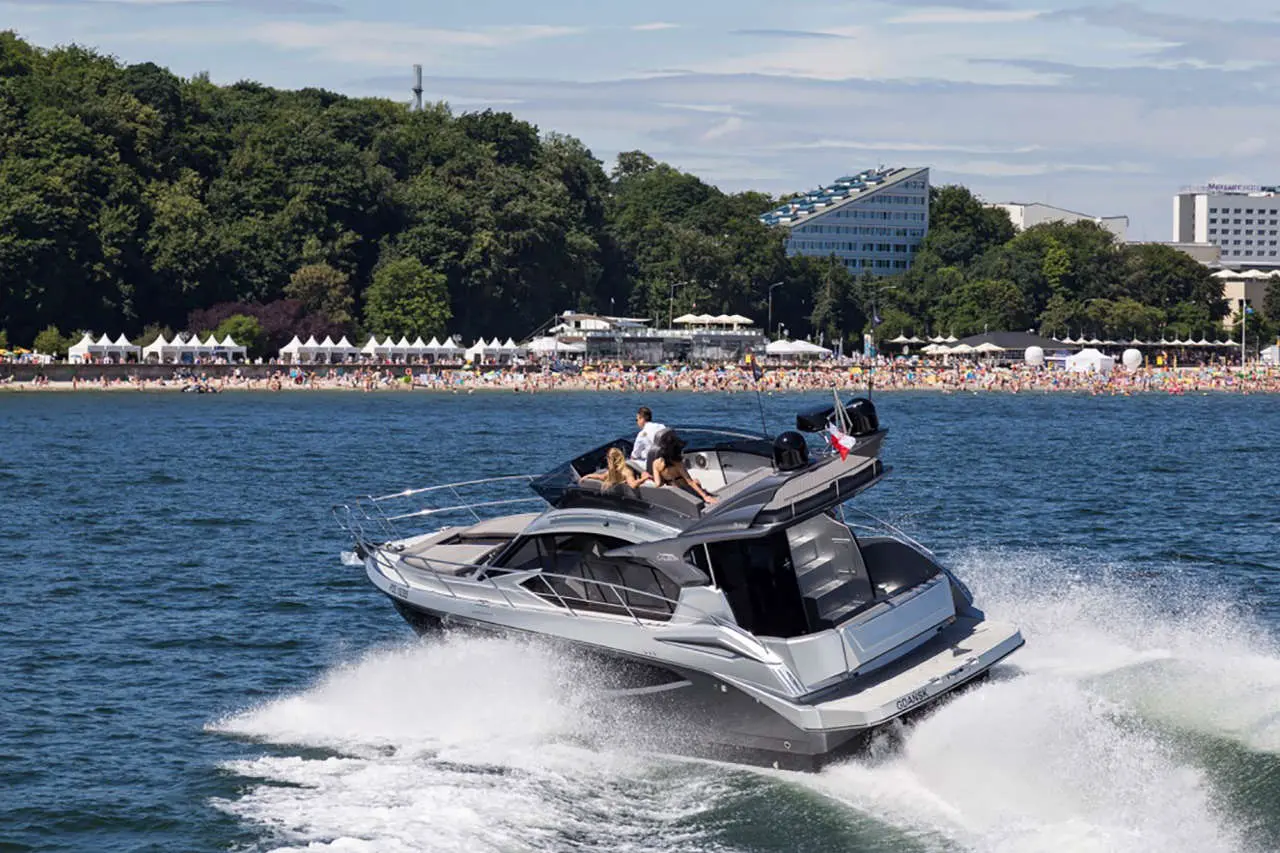 A sleek powerboat speeds across the water near a busy beach, with people and tents visible on the shore.