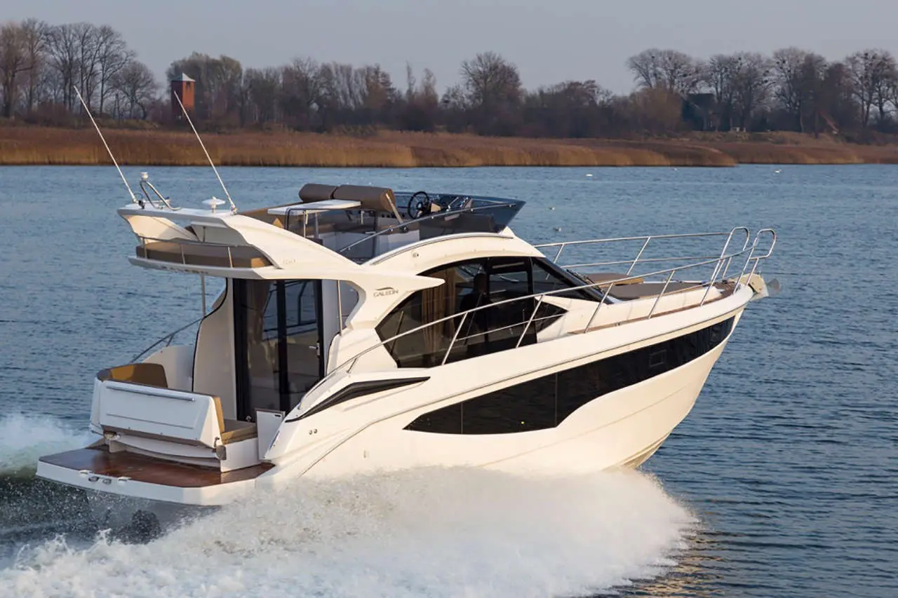 A stylish motor yacht cruising on a calm river, creating a wake, with wooded banks in the background.