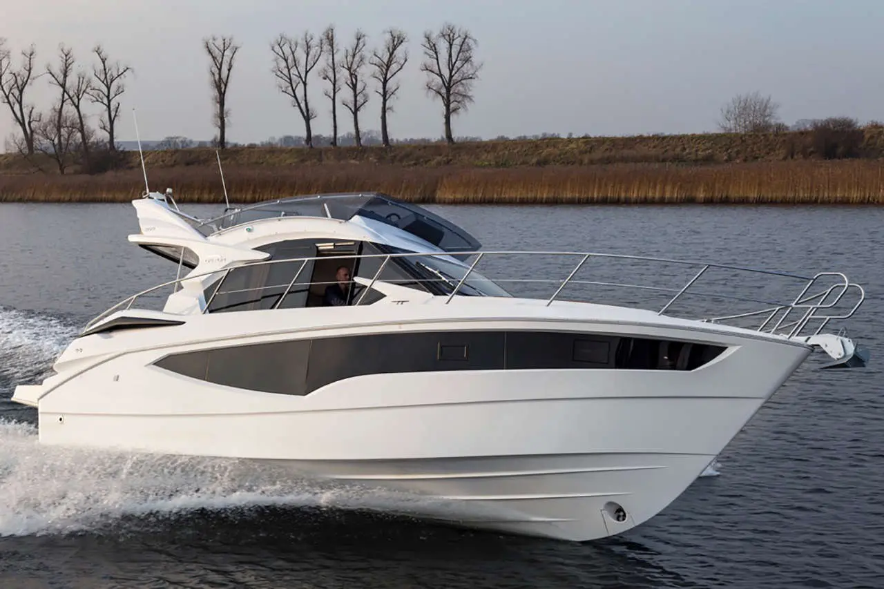 A white motorboat cuts through calm water, with a driver visible at the helm and bare trees lining the shore.