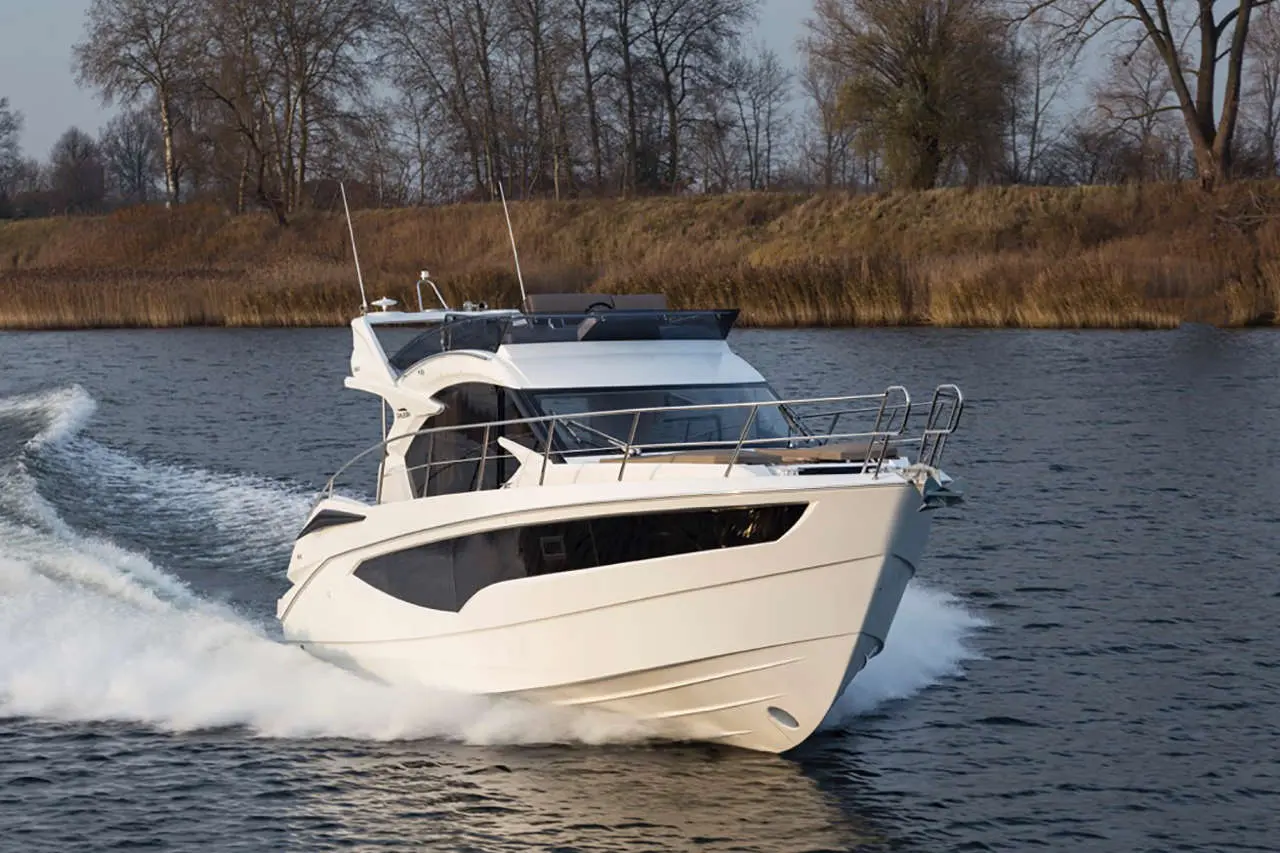 A sleek white motorboat speeds through a tranquil river, creating waves against a backdrop of trees and reeds.