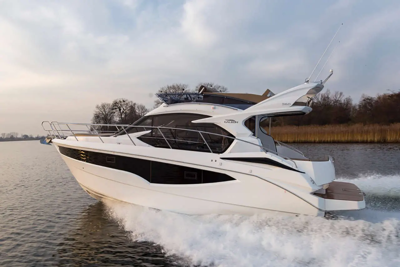 A sleek white motor yacht cruising on a calm waterway, creating a spray against a backdrop of trees and cloudy sky.