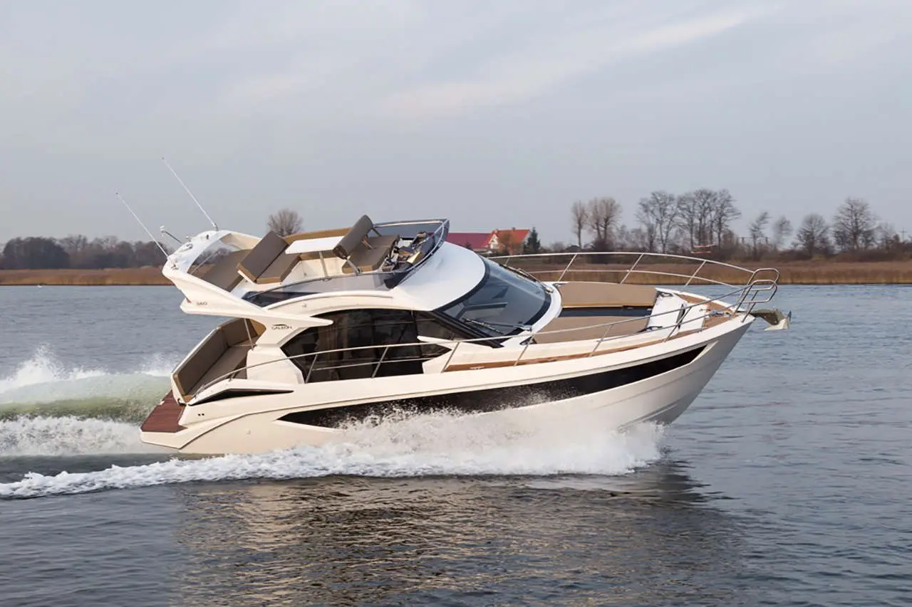 A white motorboat speeds through calm waters, creating waves, with a backdrop of trees and a distant house.