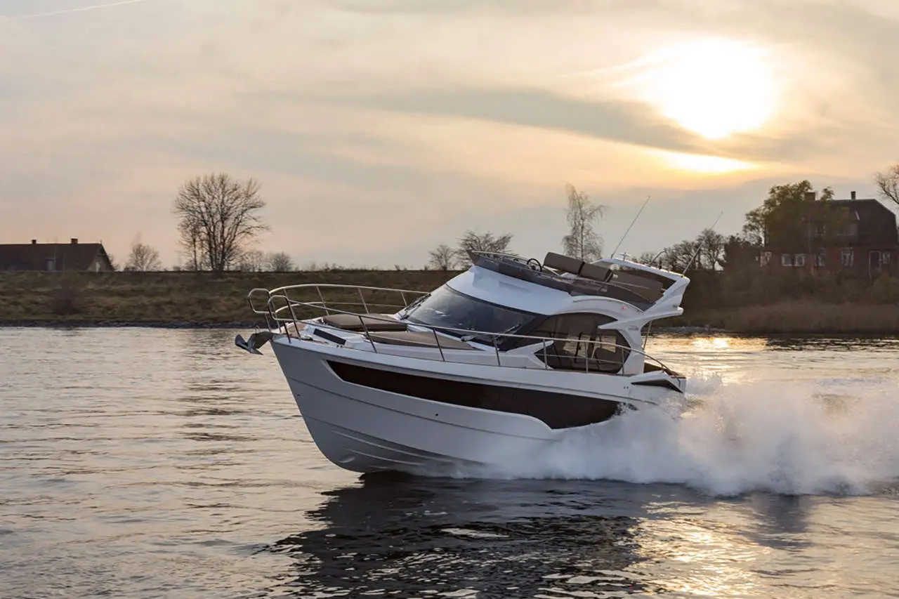 A sleek motorboat glides over water at sunset, leaving a wake, with trees and a house in the background.