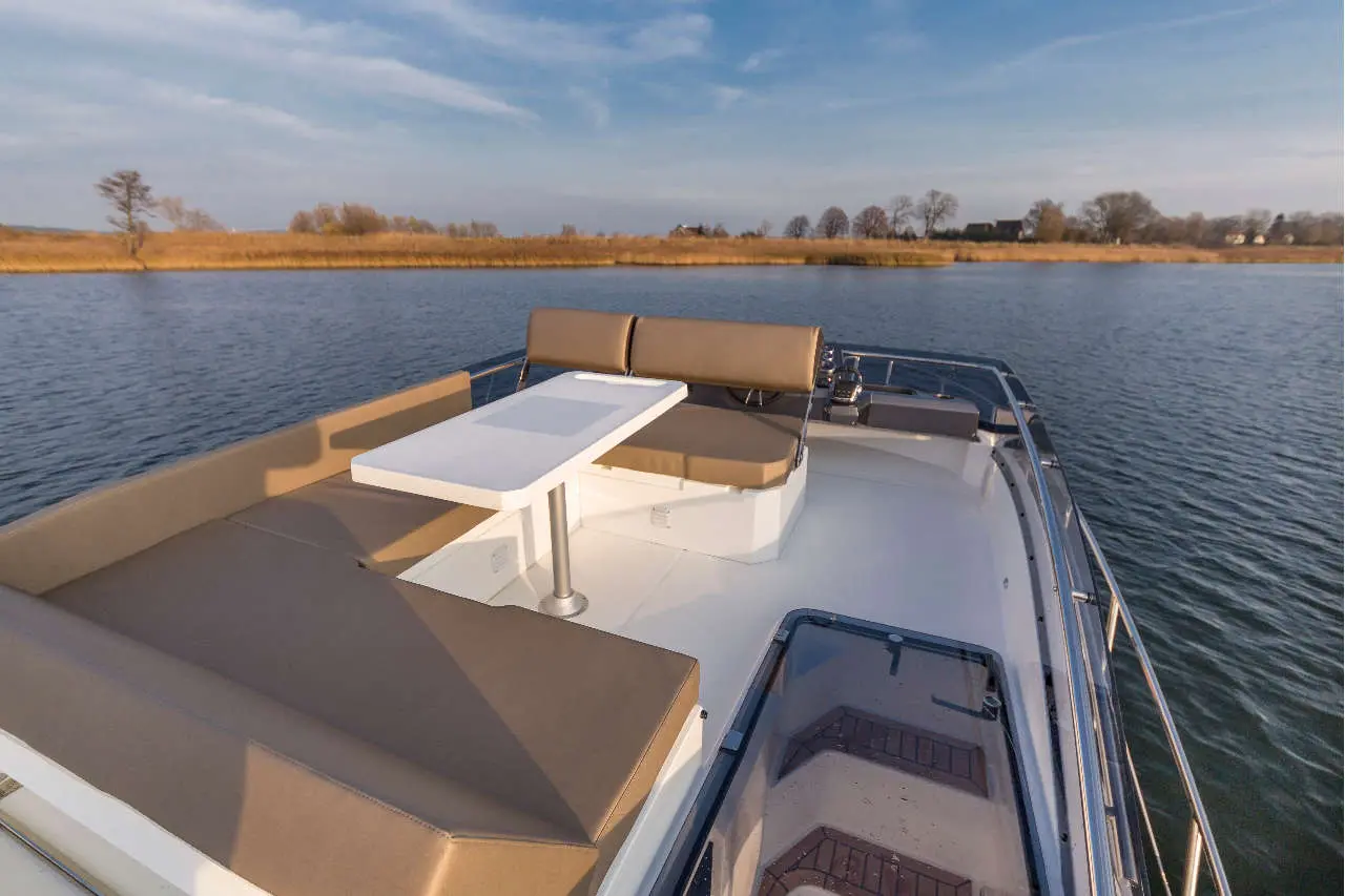 A modern boat deck featuring beige seating, a white table, and scenic views of calm water and distant trees.