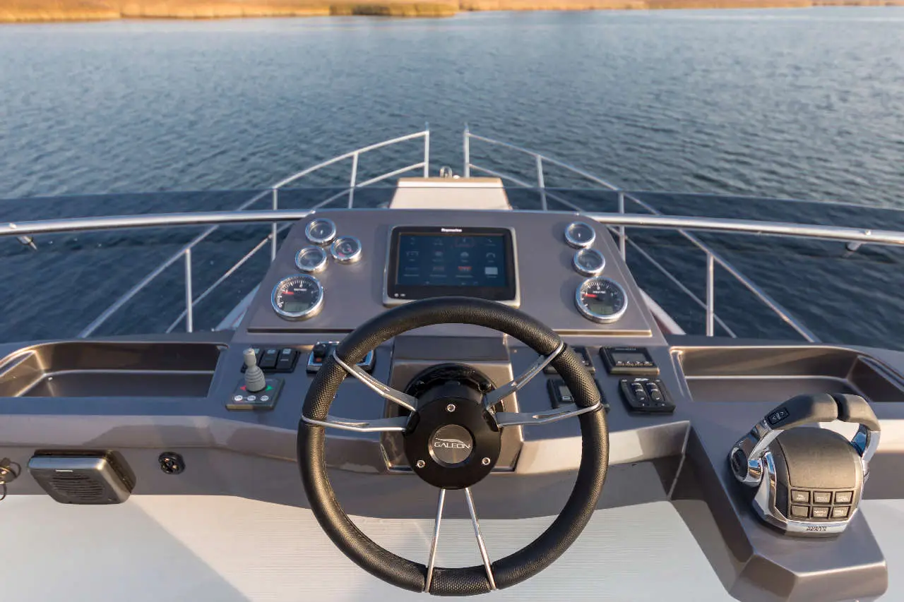 Steering wheel and controls on a yacht's cockpit, featuring a touchscreen display and multiple gauges over calm water.