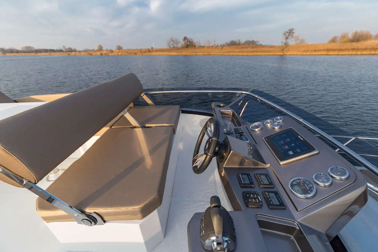 Stylish boat dashboard with steering wheel, controls, and seating, overlooking calm waters and distant shoreline vegetation.