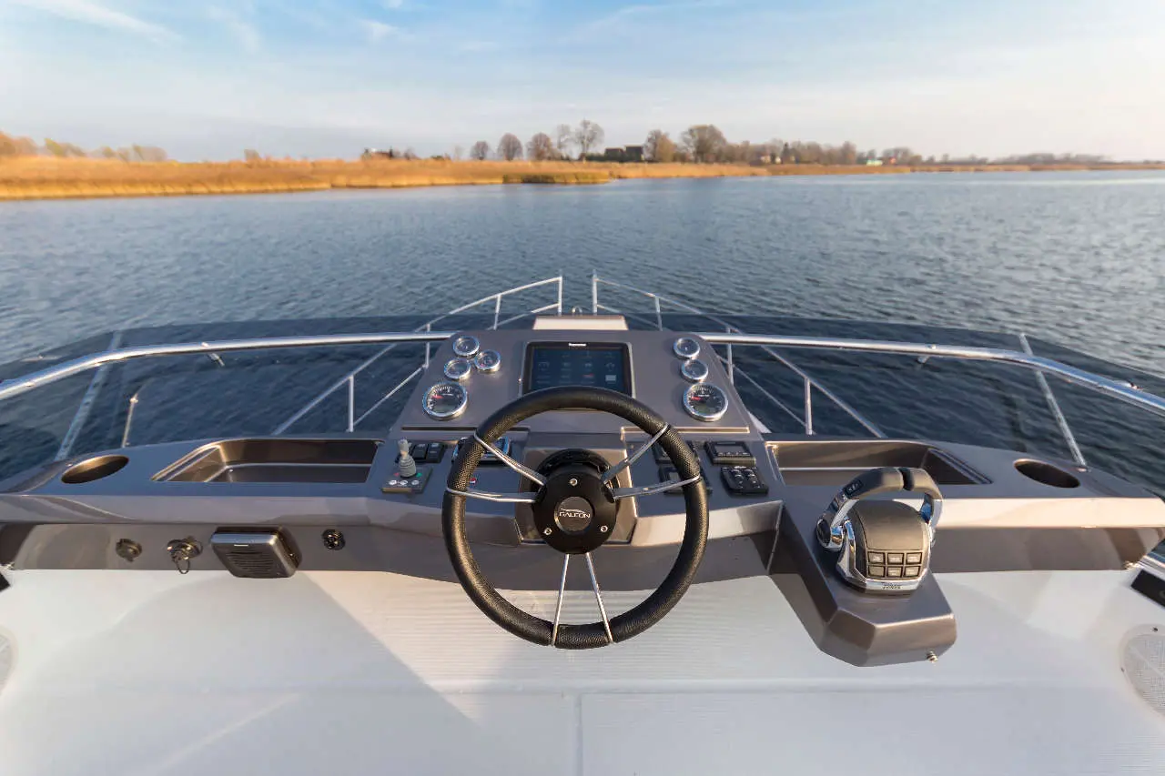 View from the steering console of a boat, featuring a steering wheel and control panel against a serene water backdrop.