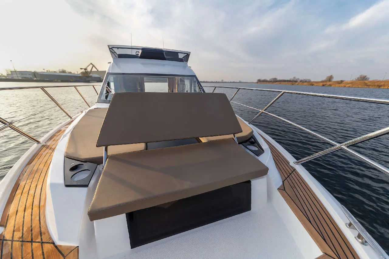 Luxury motorboat deck with cushioned seating, overlooking calm water and distant industrial landscape under a cloudy sky.