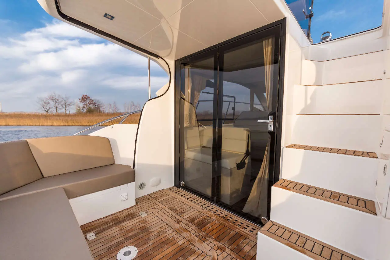 A luxury boat deck with a plush seating area, glass door entrance, and stairs leading to the upper deck, overlooking water.