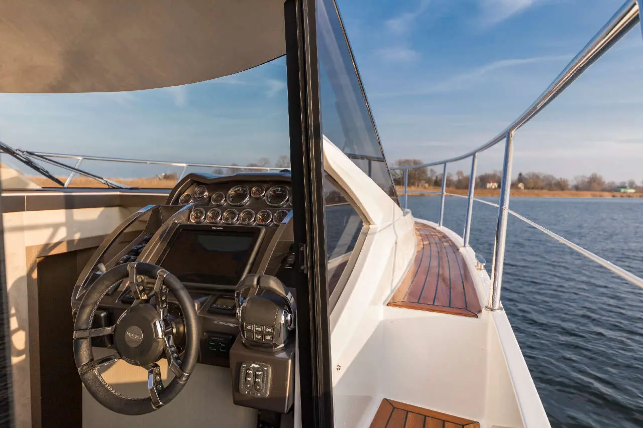 Luxury boat interior showing the steering wheel and control console, with water and shoreline visible through the window.
