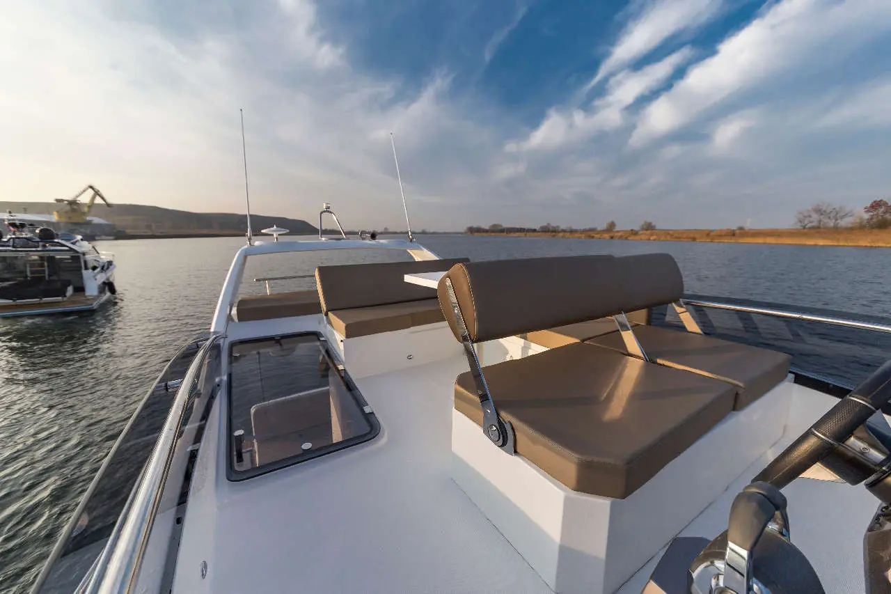 A boat's deck features cushioned seating, with calm water and a scenic background under a blue sky with clouds.