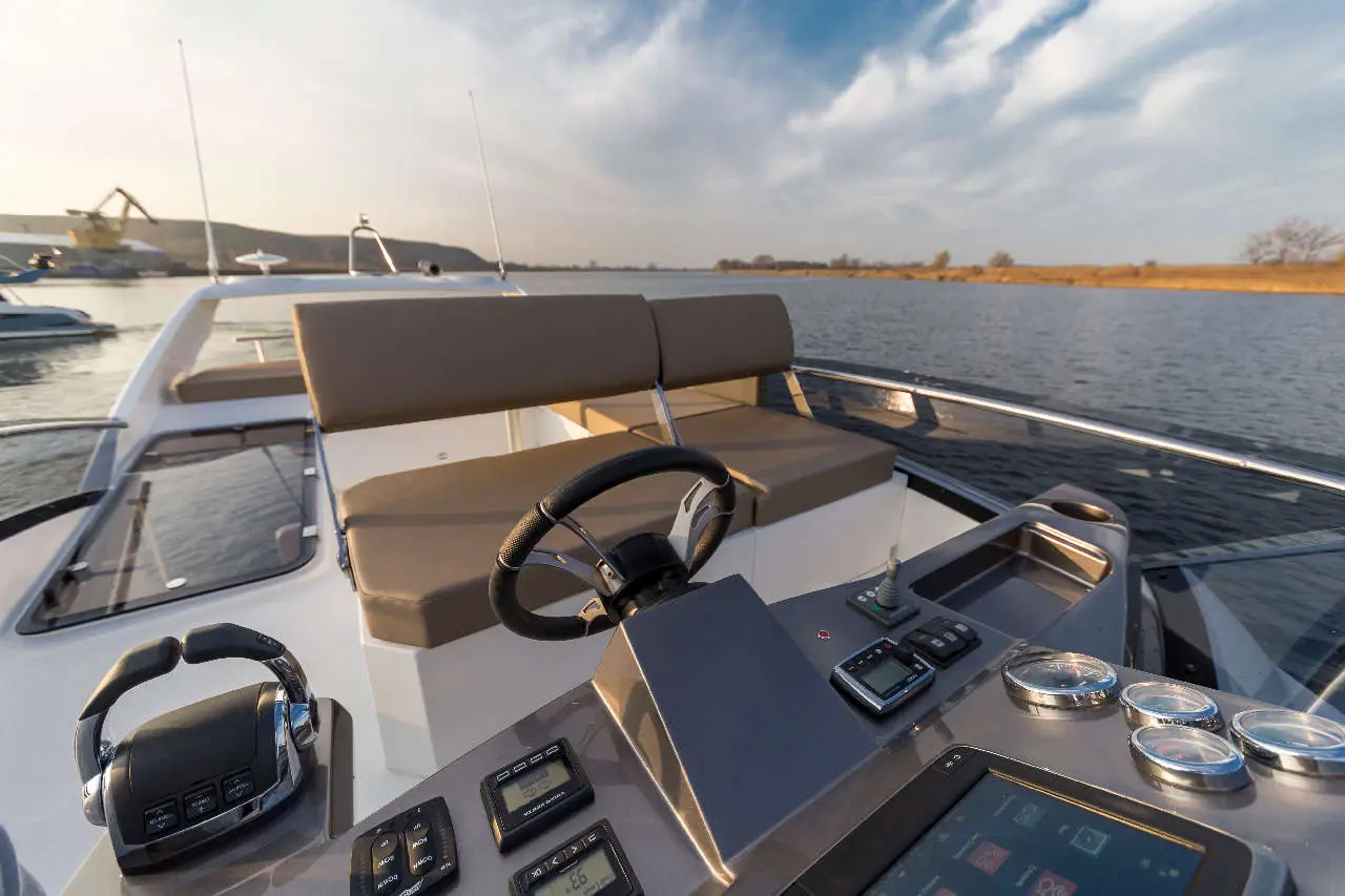 A luxury boat dashboard with a steering wheel and navigation controls, overlooking calm waters and scenic landscape.